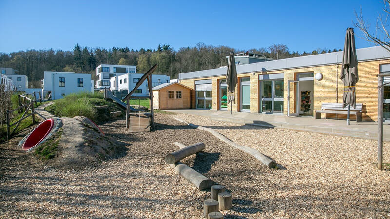 Ein moderner Spielplatz mit Rutsche, Klettergerüst und Holzhütte befindet sich vor einem flachen, einstöckigen Gebäude, umgeben von Bäumen und mehrstöckigen weißen Wohnhäusern.