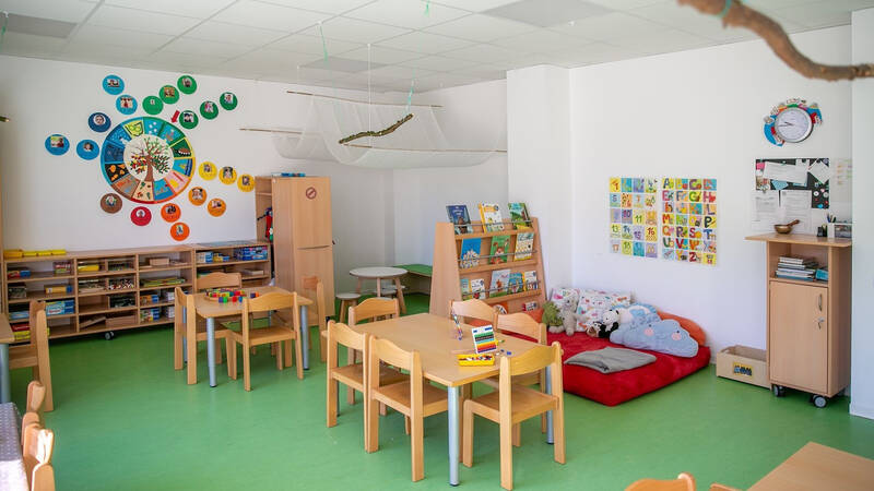 Ein farbenfrohes Kinderzimmer mit mehreren kleinen Holztischen und Stühlen, Regalen mit Büchern und Spielsachen, einer Kuschelecke und Wanddekorationen mit Buchstaben und Zahlen.