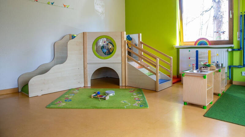 Ein heller Kindergartenraum mit grünem Teppich, einer kleinen Holzrutsche, einer Kinderspielküche und einem Fenster, das Tageslicht hereinlässt.
