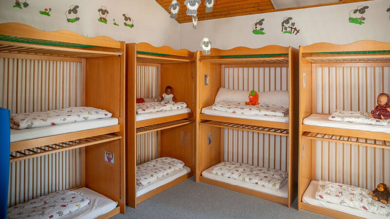 Ein Kindergarten-Schlafraum mit mehreren Holz-Etagenbetten, in denen Stofftiere und Puppen liegen, umgeben von Wänden mit Schafmotiven.