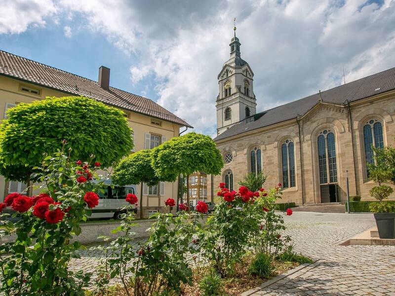 Auf dem Bild ist ein Kirchplatz mit gepflastertem Boden zu sehen, umgeben von Gebäude und grünen Bäumen sowie blühenden roten Rosen, vor einem klaren Himmel.
